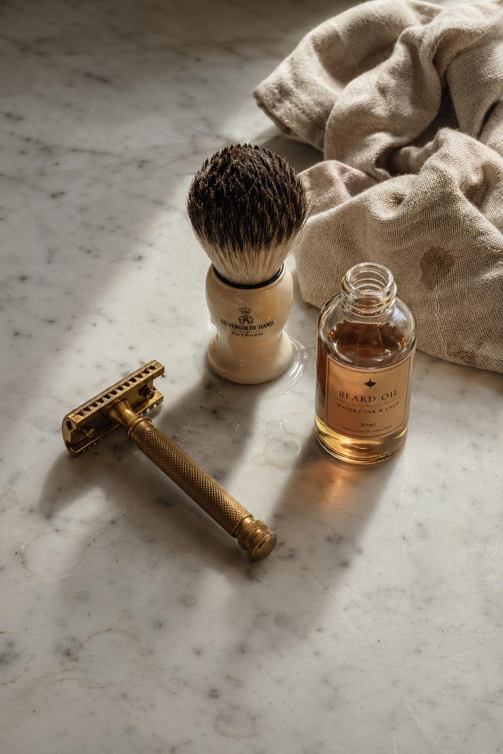 A warm still life on marble: a polished brass safety razor, badger-hair shaving brush, and an apothecary bottle of amber oil with rising steam.